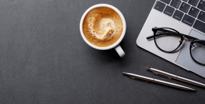 This image features a black background with a white coffee mug; the coffee is visible inside next to the coffee cup is two black pens at the top right of the image there is the edge of a laptop visible with glasses on the keyboard.
