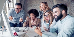 Diverse group of people at work huddled around a desk covered in papers celebrating with a brick wall background