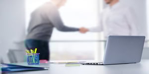 Focused desk with cupholder and computer layered on top of blurred businessmen shaking hands