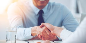 Man in light blue button up shirt and navy tie, shaking hands with person in white long sleeve shirt over top of desk with documents on it