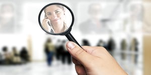 Woman's reflection smiling in a magnifier with her hand holding magnifier, on top of blurred office background with small crowd