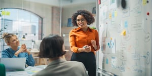 Two women sitting at desk at work looking at woman presenting on a white board