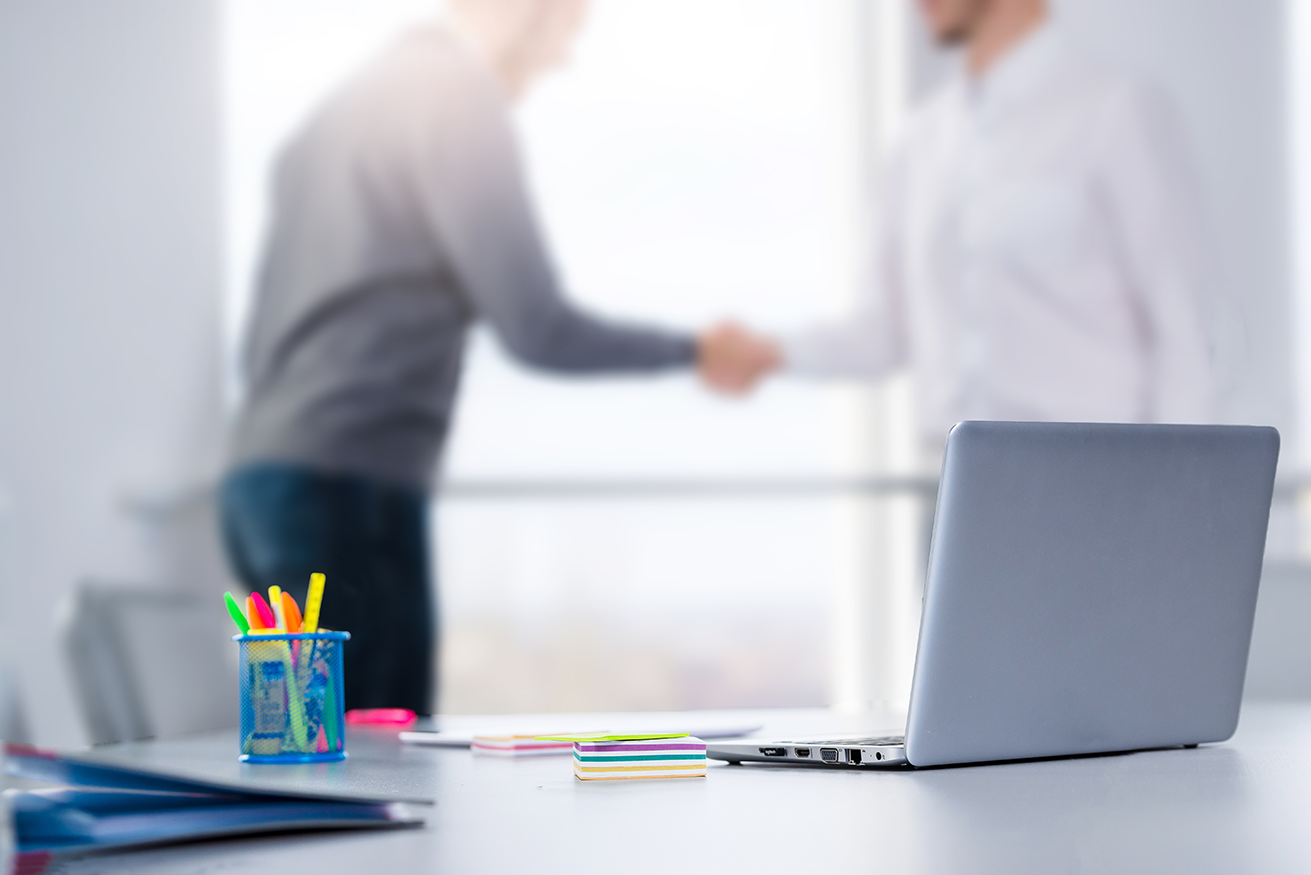 Focused desk with cupholder and computer layered on top of blurred businessmen shaking hands