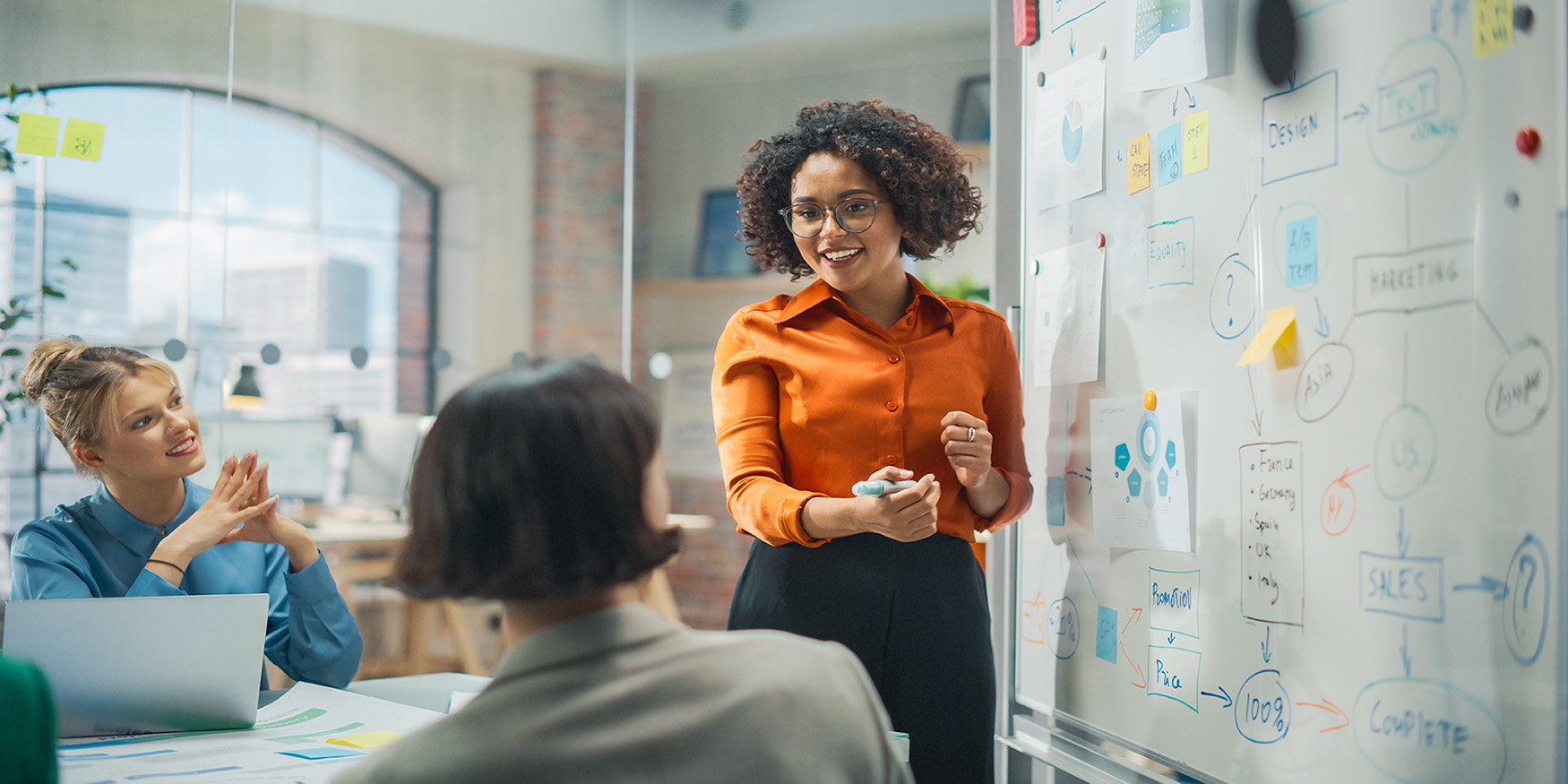 Two women sitting at desk at work looking at woman presenting on a white board